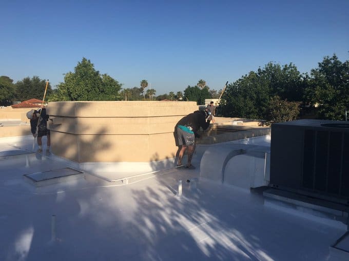 Workers applying a white coating to a flat roof with parapet walls, showing a maintenance process.